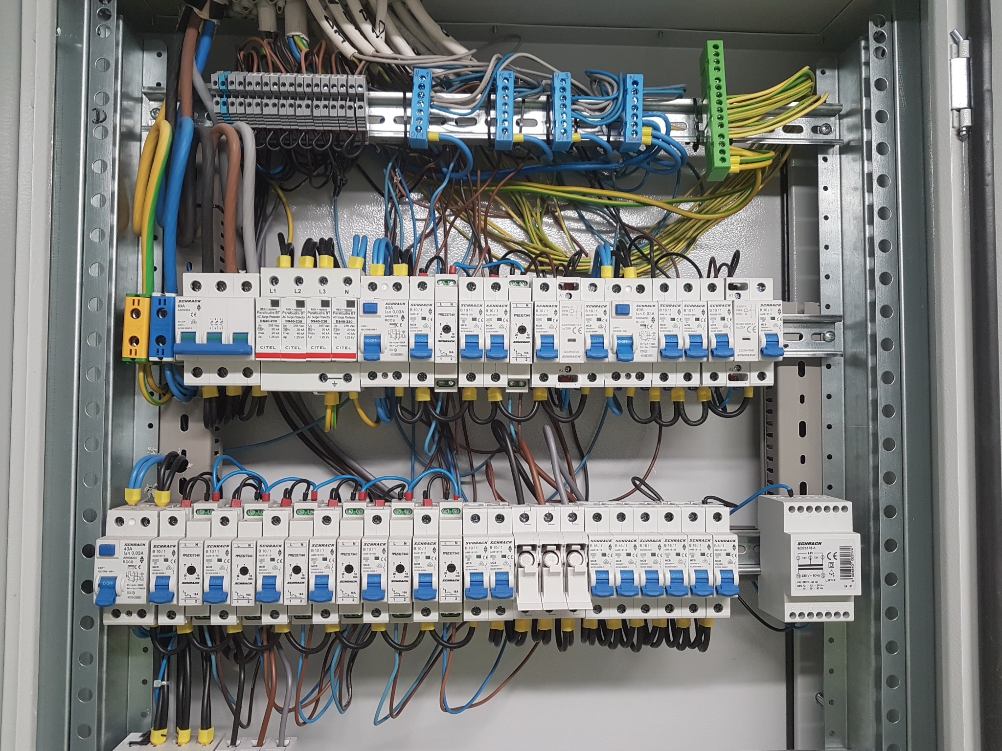 Rozdzielnica elektryczna z zainstalowanymi bezpiecznikami, zabezpieczeniami przeciwprzepięciowymi i przewodami o różnych kolorach, widoczne oznaczenia na urządzeniach.