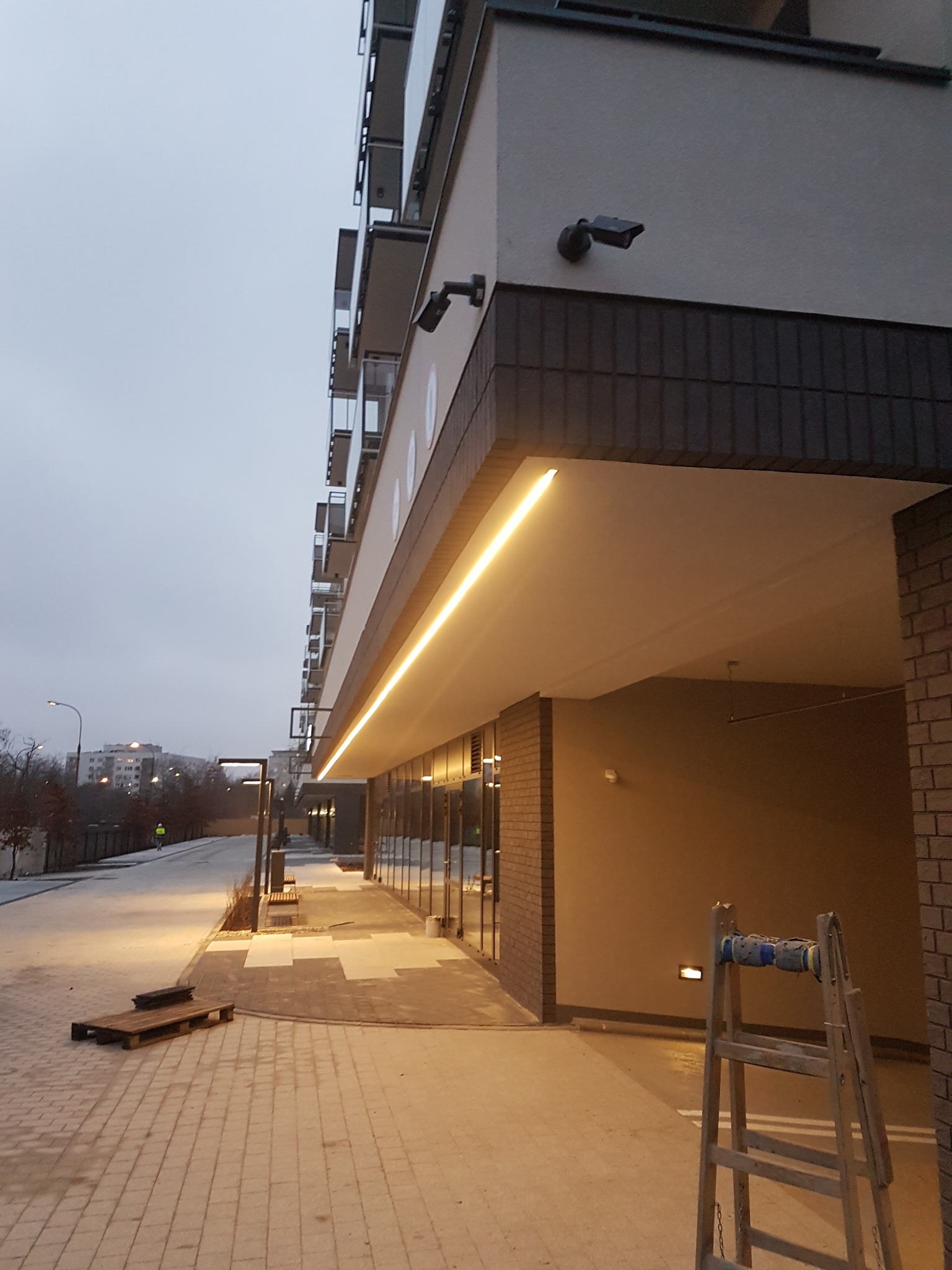 Nowoczesny budynek z podświetlanym LED-owo podcieniem, widoczna drabina z niebieskimi elementami na szczycie, kamera monitoringu, bruk na zewnątrz i elementy małej architektury w tle.