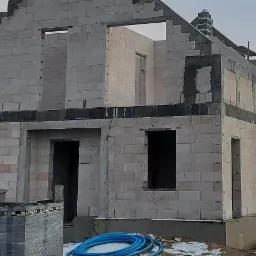 Surowy stan domu z bloczków, widoczne otwory okienne i drzwiowe, częściowo ośnieżony teren, paleta bloczków LEWTAK i zwój niebieskiego węża na pierwszym planie.