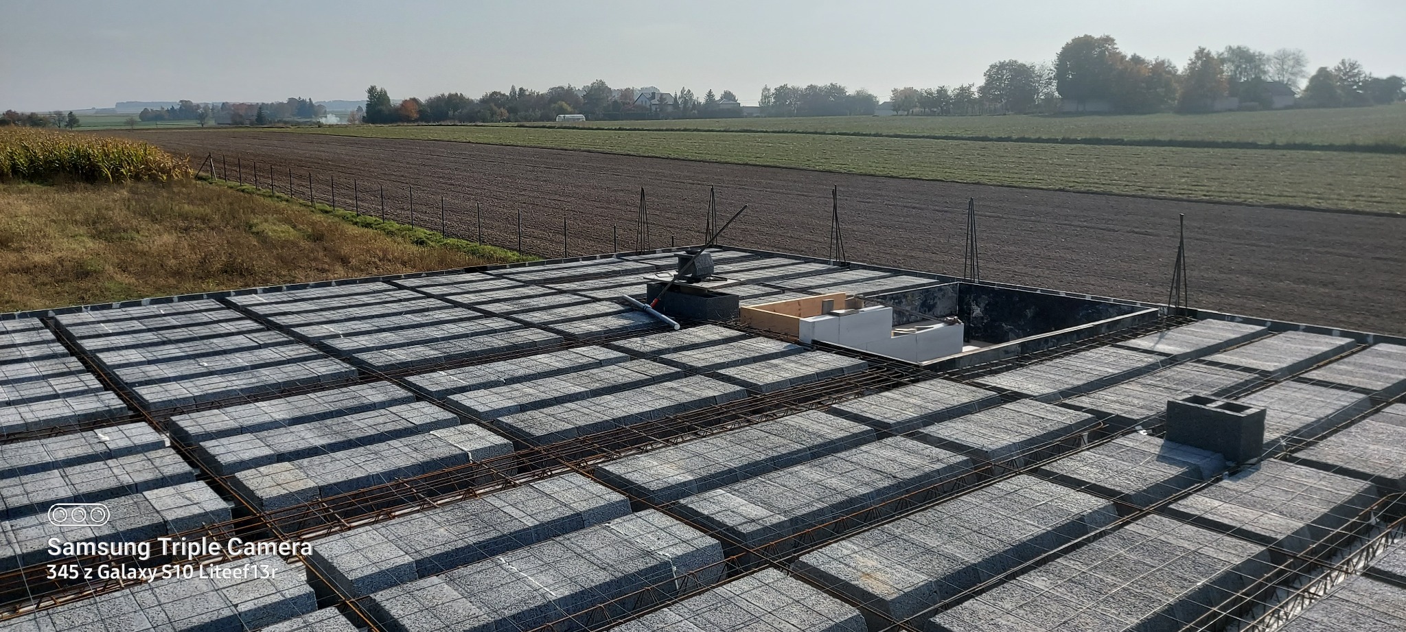 Wykonywanie stropu Teriva z widocznymi bloczkami betonowymi i zbrojeniem stalowym, na tle pola uprawnego i zabudowań wiejskich w oddali, w słoneczny dzień.