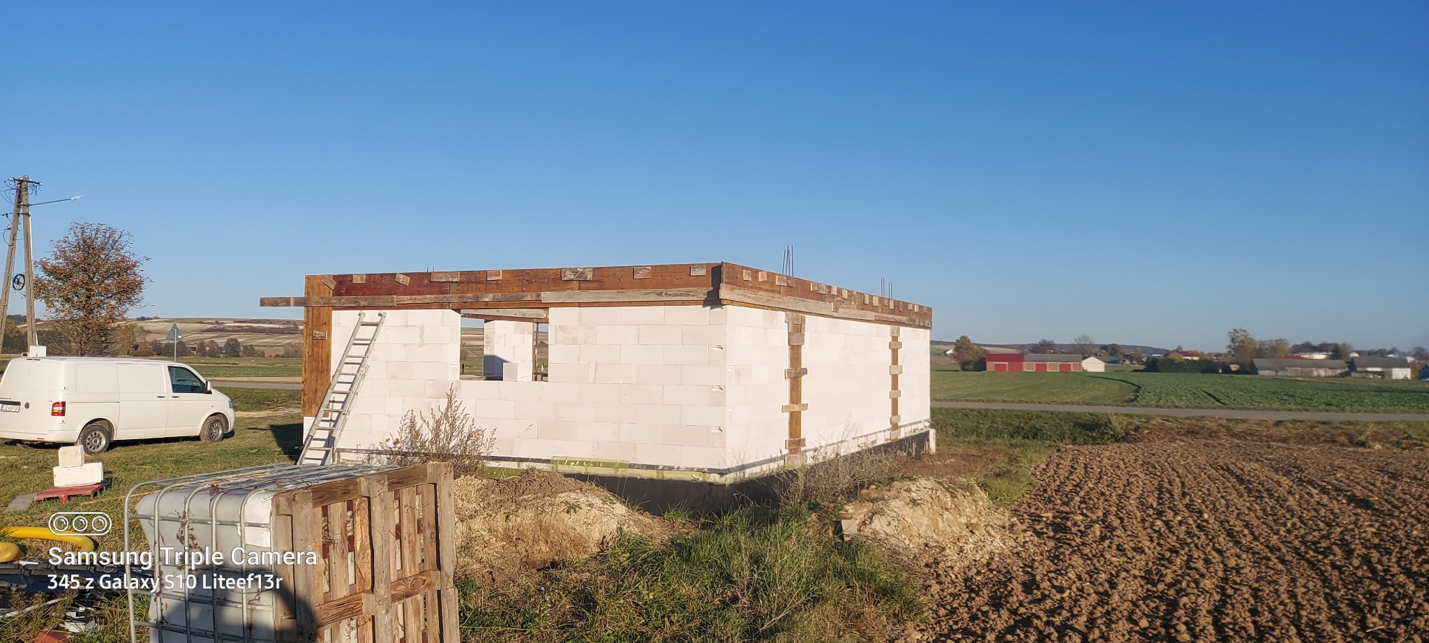 Wznoszenie ścian parterowego domu z białych bloczków, widoczne deskowanie stropu, drabina oparta o ścianę, w tle pola uprawne i błękitne niebo.