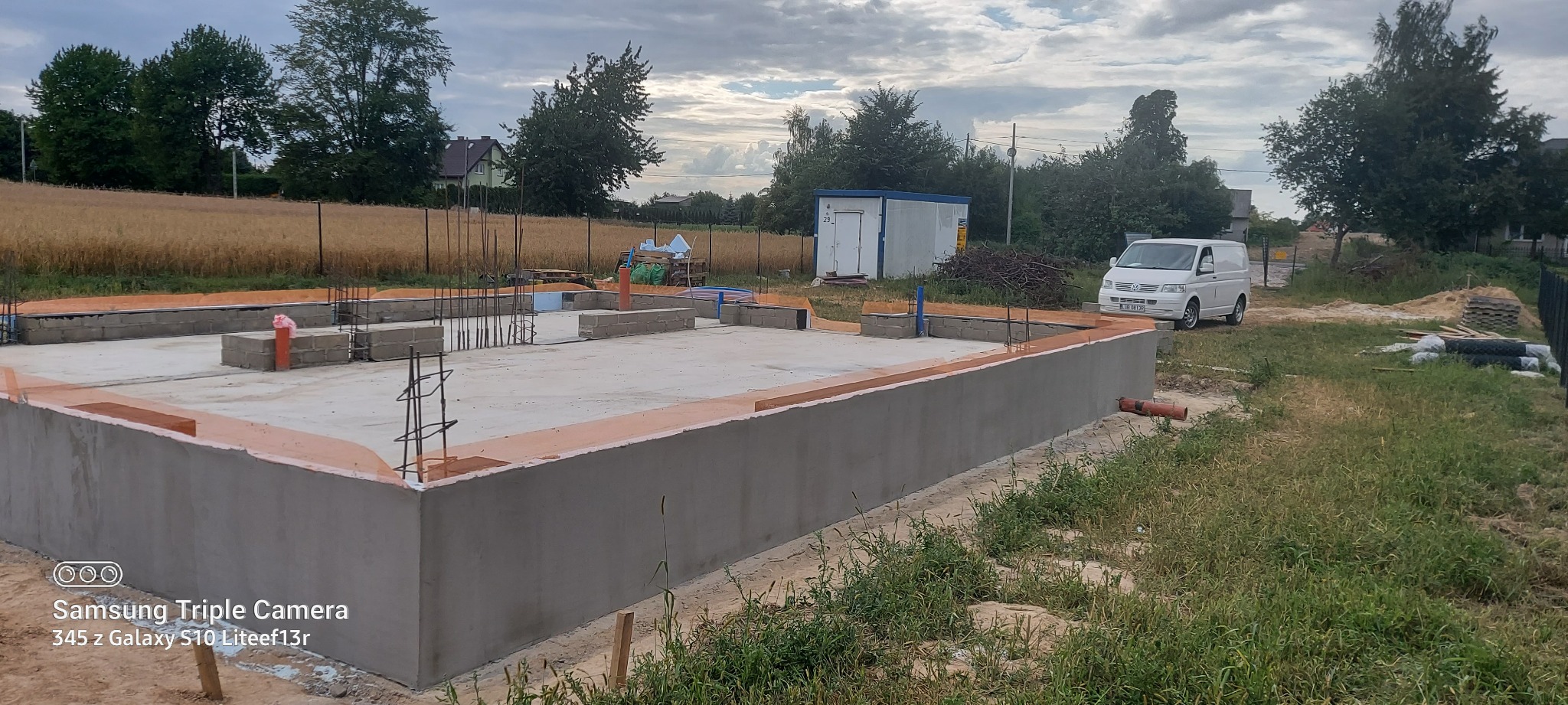 Wylany fundament betonowy z izolacją termiczną, widoczne zbrojenia i bloczki fundamentowe, na działce budowlanej z zaparkowanym białym vanem w tle.