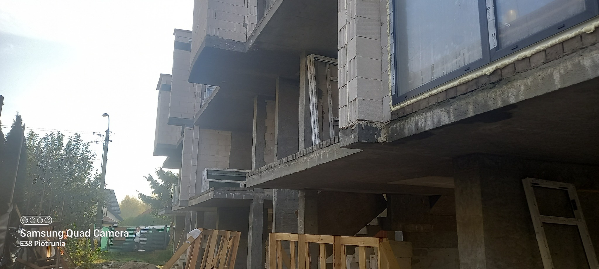 Surowy stan budynku wielorodzinnego w trakcie wznoszenia, widoczne bloczki betonowe, zamontowane okna z pianką montażową, drewniane ramy okienne i palety na pierwszym planie.