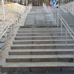 Betonowe schody zewnętrzne z nierdzewnymi poręczami, prowadzące w górę zbocza, widoczne osoby w oddali.