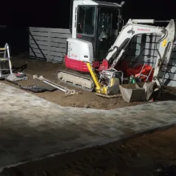 Nocne ujęcie częściowo ułożonej ścieżki z kostki brukowej w kształcie litery L, z minikoparką Takeuchi TB23R, zagęszczarką, łopatą i drabiną w tle, przy betonowym ogrodzeniu.