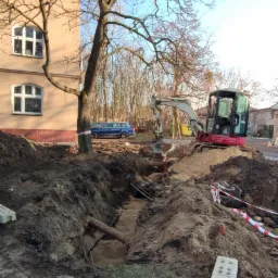 Wykopany rów z widocznymi rurami, minikoparka na skraju wykopu, budynek mieszkalny w tle, zabezpieczenia taśmą ostrzegawczą.