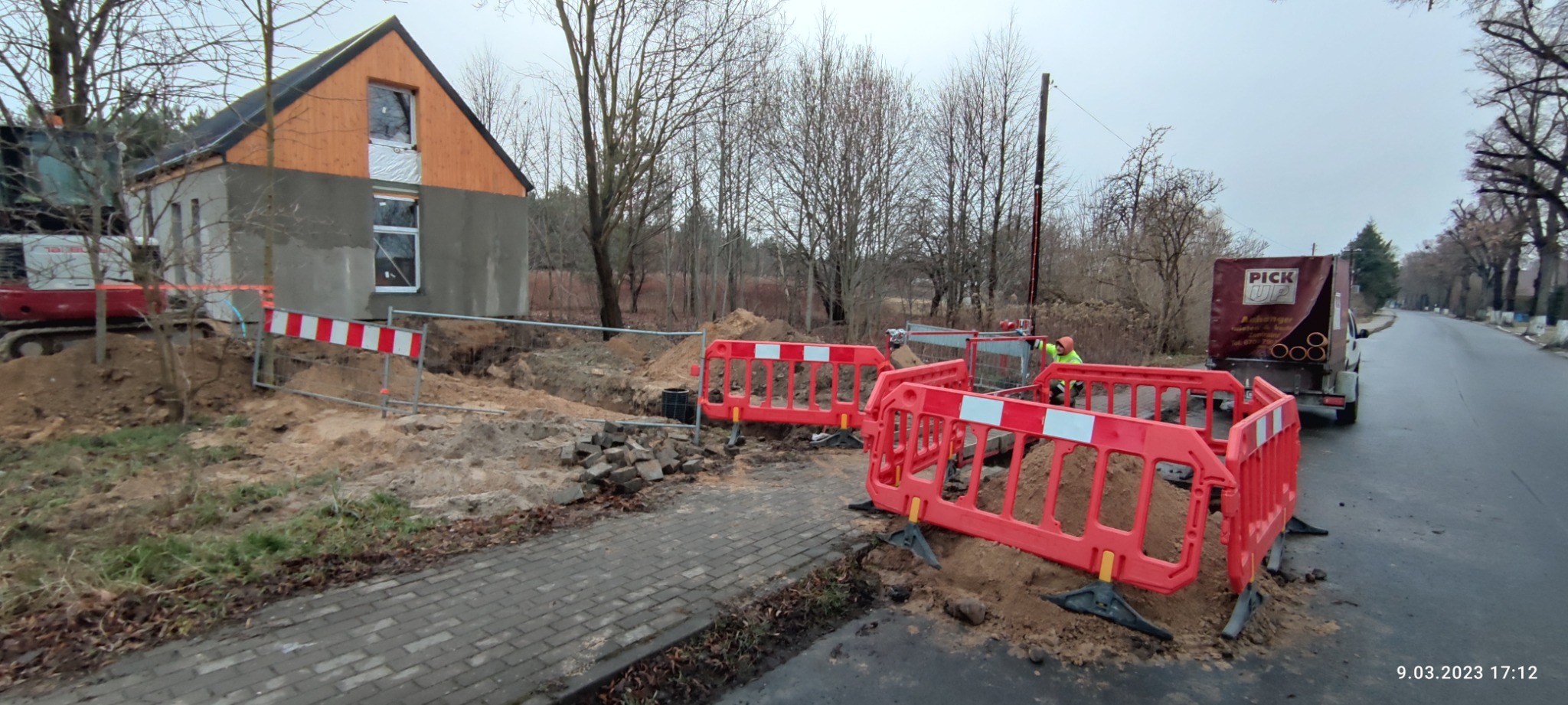 Wykonanie wykopu pod przyłącze wodno-kanalizacyjne przy budowie domu jednorodzinnego, zabezpieczone barierami ochronnymi, widoczny mini-koparka i samochód dostawczy z rurami.