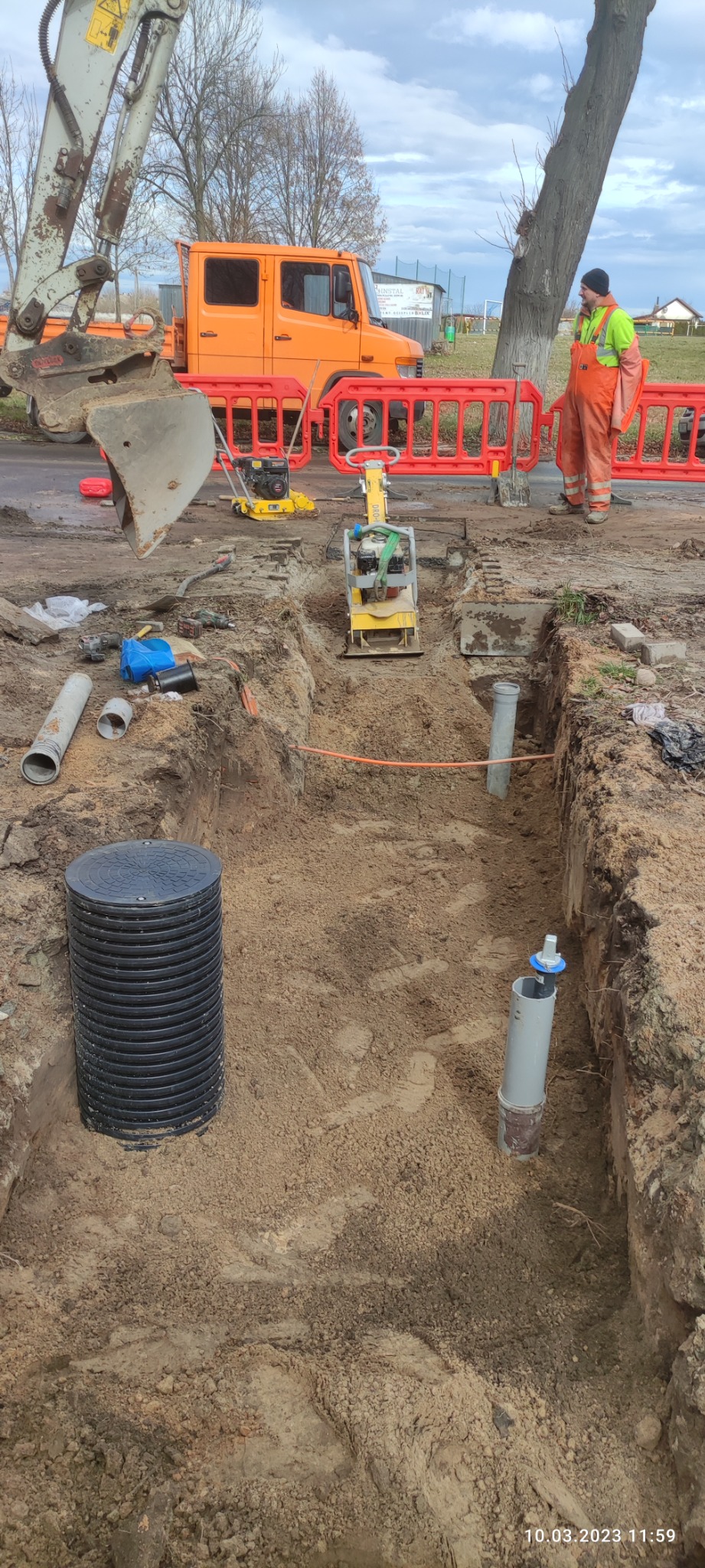 Wykop w ziemi z widocznymi rurami kanalizacyjnymi, studzienką rewizyjną, zagęszczarką i pomarańczową ciężarówką w tle, zabezpieczony barierkami i nadzorowany przez pracownika w kombinezonie.