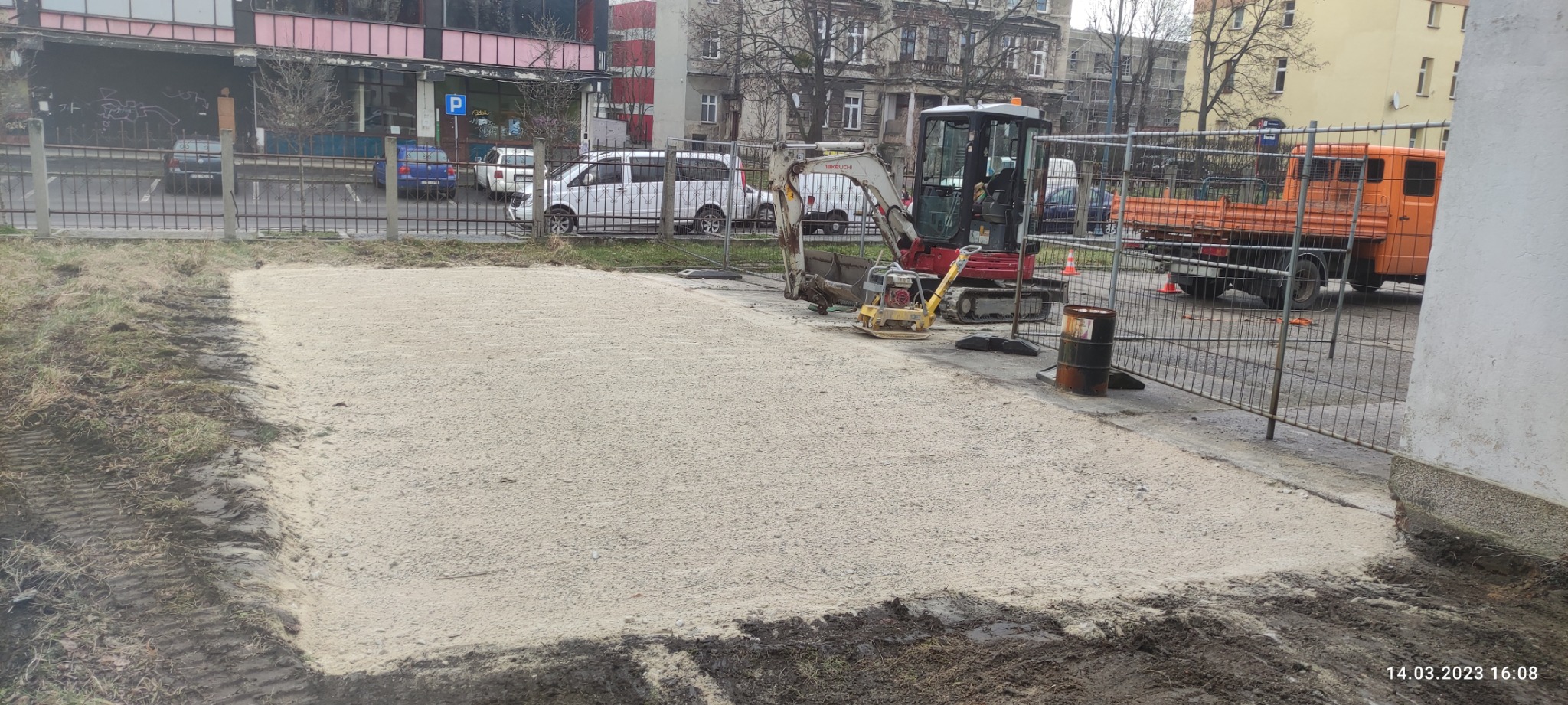 Przygotowany, wyrównany i utwardzony teren pod budowę, widoczny minikoparka, zagęszczarka wibracyjna, pomarańczowa ciężarówka i metalowe ogrodzenie tymczasowe.