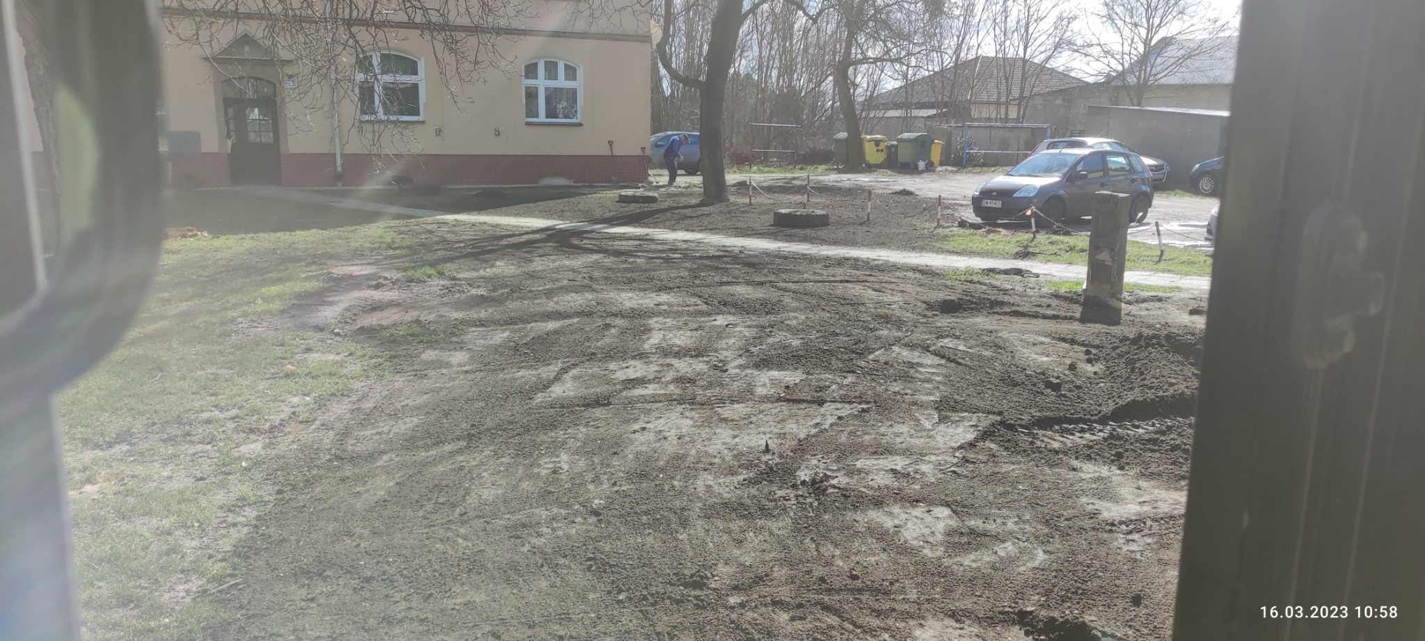 Przygotowany teren pod budowę z widocznymi śladami opon, budynek w tle, częściowo zasłonięty widok z kabiny maszyny.