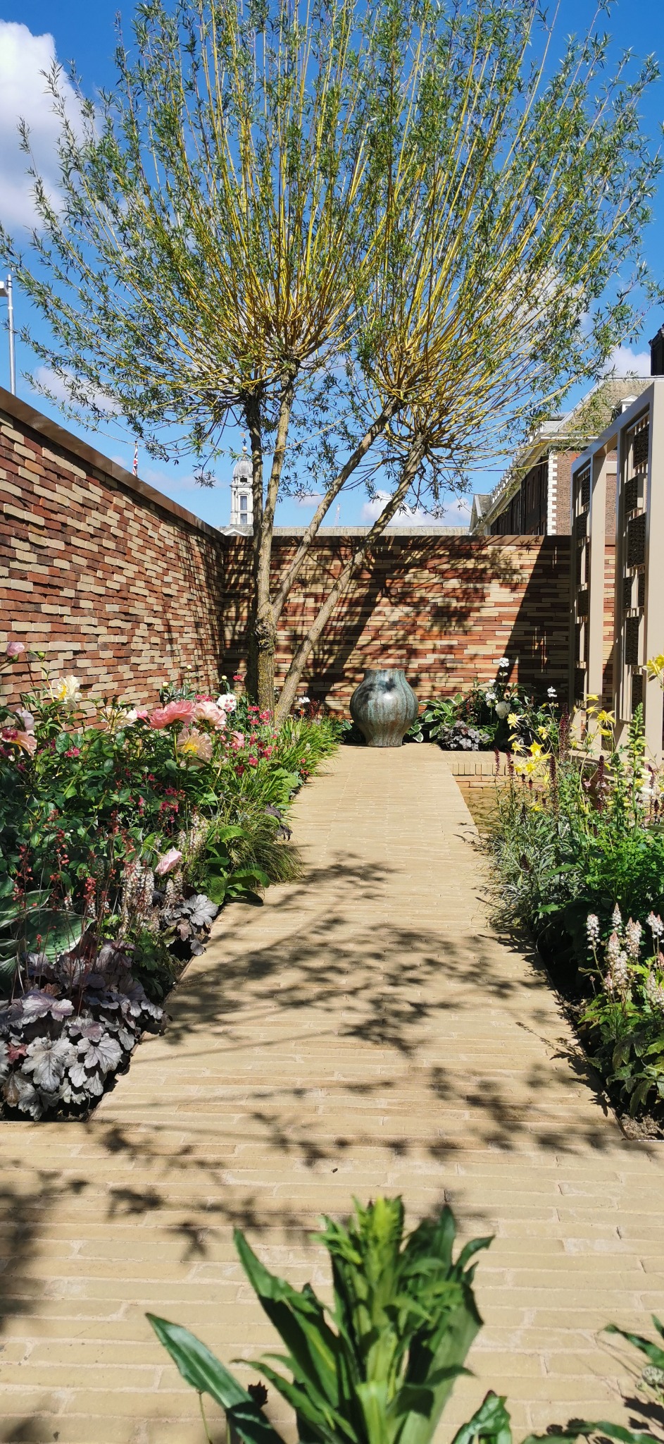 Urokliwy ogród z ceglaną ścieżką, rabatami kwiatowymi i drzewem o żółtych gałęziach, zakończony ozdobną ceramiczną wazą na tle ceglanego muru.