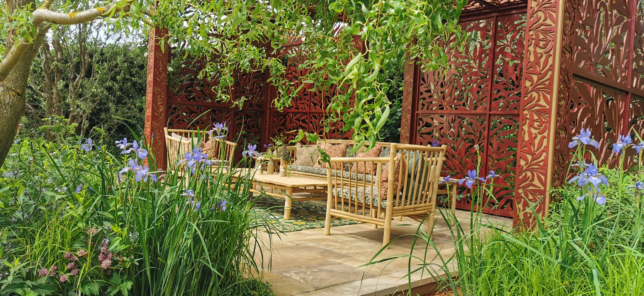 Zadaszona altana z ażurowymi, metalowymi ścianami, bambusowe meble ogrodowe z poduszkami, irysy syberyjskie na pierwszym planie, kamienna podłoga.