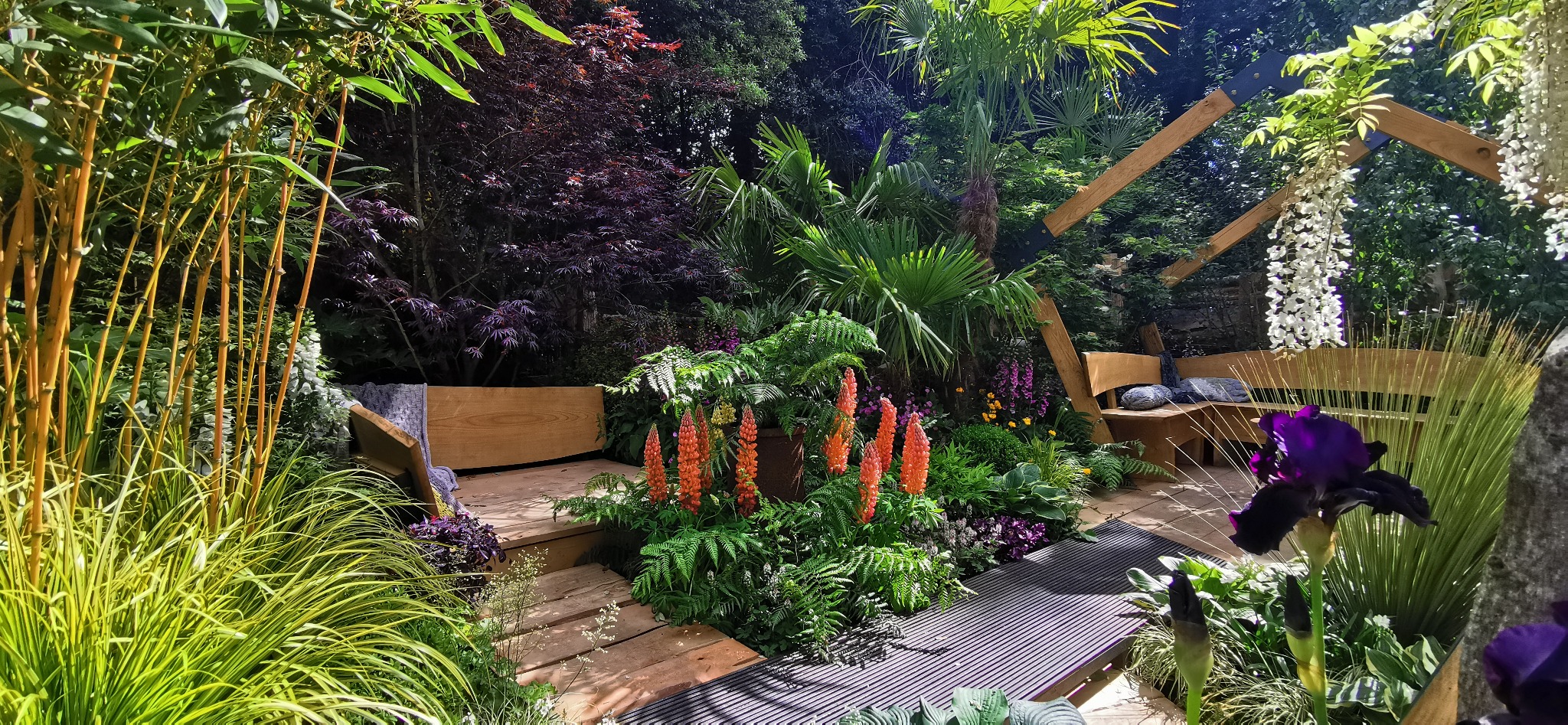 Bogato obsadzony ogród w Rudzińcu z drewnianym tarasem, egzotycznymi roślinami, kwitnącymi pomarańczowymi knifofiami i bambusami, tworzący zaciszne miejsce do relaksu.