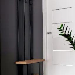 Nowoczesny wieszak na ubrania z metalową konstrukcją i drewnianymi półkami, zamontowany na ciemnej ścianie w przedpokoju, obok białych drzwi z poziomymi frezowaniami.