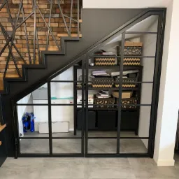 Metalowa konstrukcja schodów z ażurową balustradą i przeszklonymi drzwiami do przestrzeni pod schodami, z widoczną ceglaną ścianą w tle.