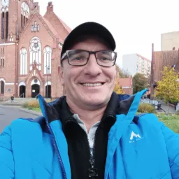 Uśmiechnięty mężczyzna w okularach i niebieskiej kurtce McKinley na tle ceglanego kościoła z wieżą zegarową i ulicy.