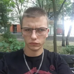 Młody mężczyzna w okularach i czarnej koszulce z nadrukiem, siedzący na ławce w parku, z budynkiem w tle.