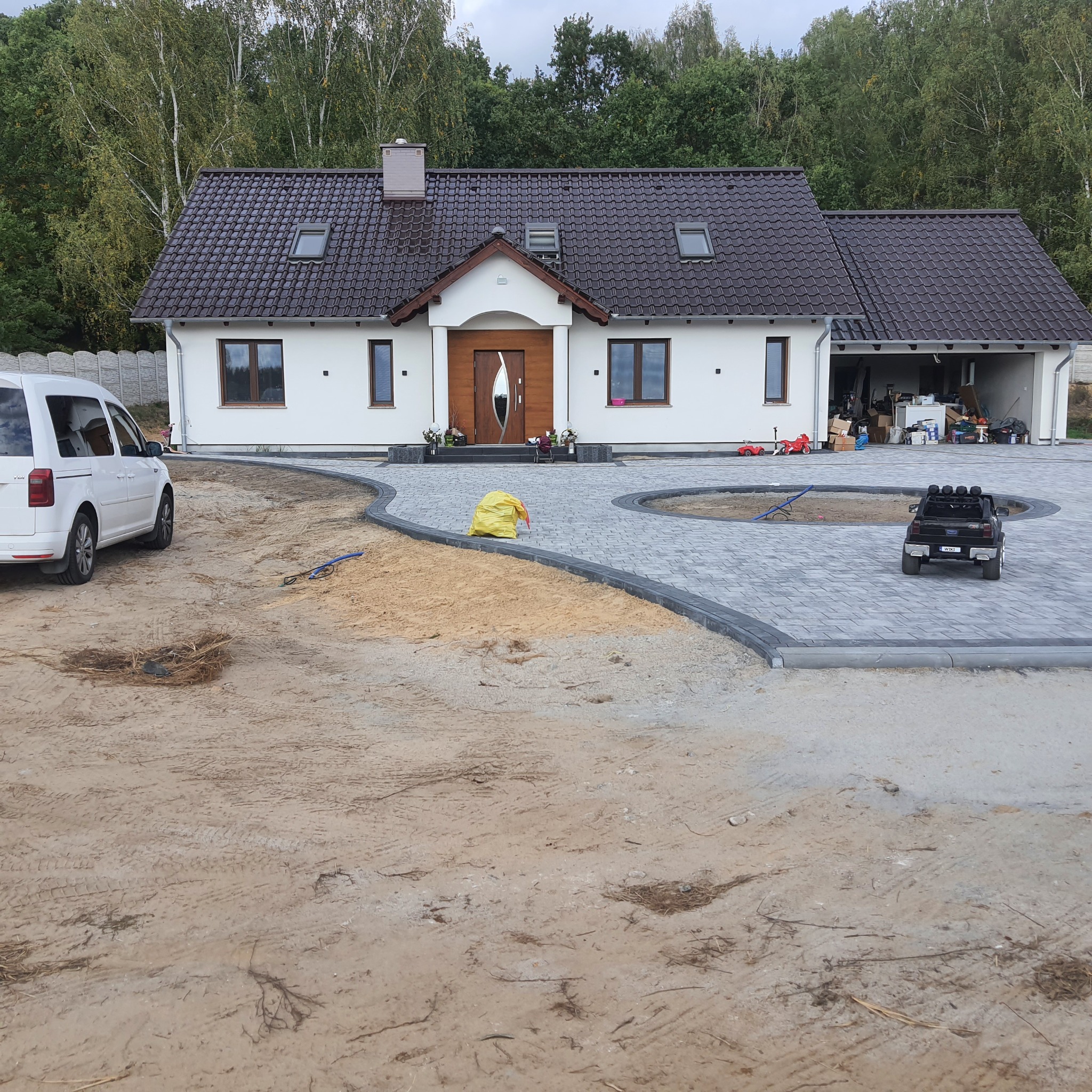 Nowo wybrukowany podjazd z szarej kostki, z okrągłym wzorem i ciemnoszarymi obrzeżami, prowadzący do białego domu z brązowym dachem, na podwórku stoi biały van i czarny samochód-zabawka.
