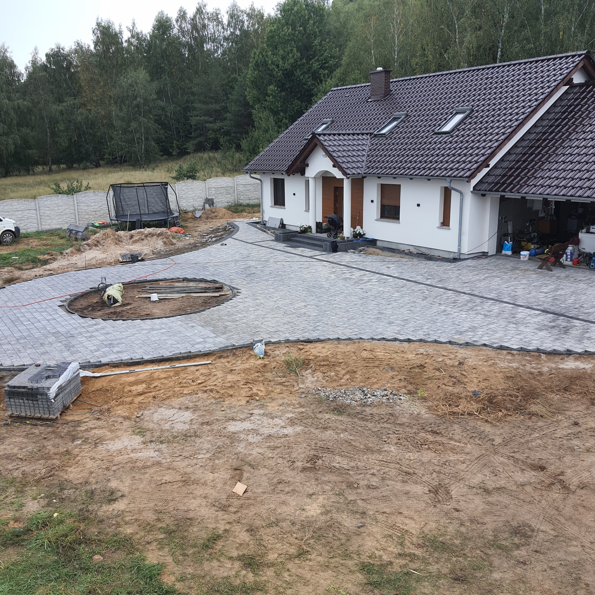 Ułożona kostka brukowa na podjeździe domu jednorodzinnego, widoczne prace wykończeniowe wokół okrągłego klombu, niezakończone krawężniki, paleta z kostką w rogu zdjęcia.