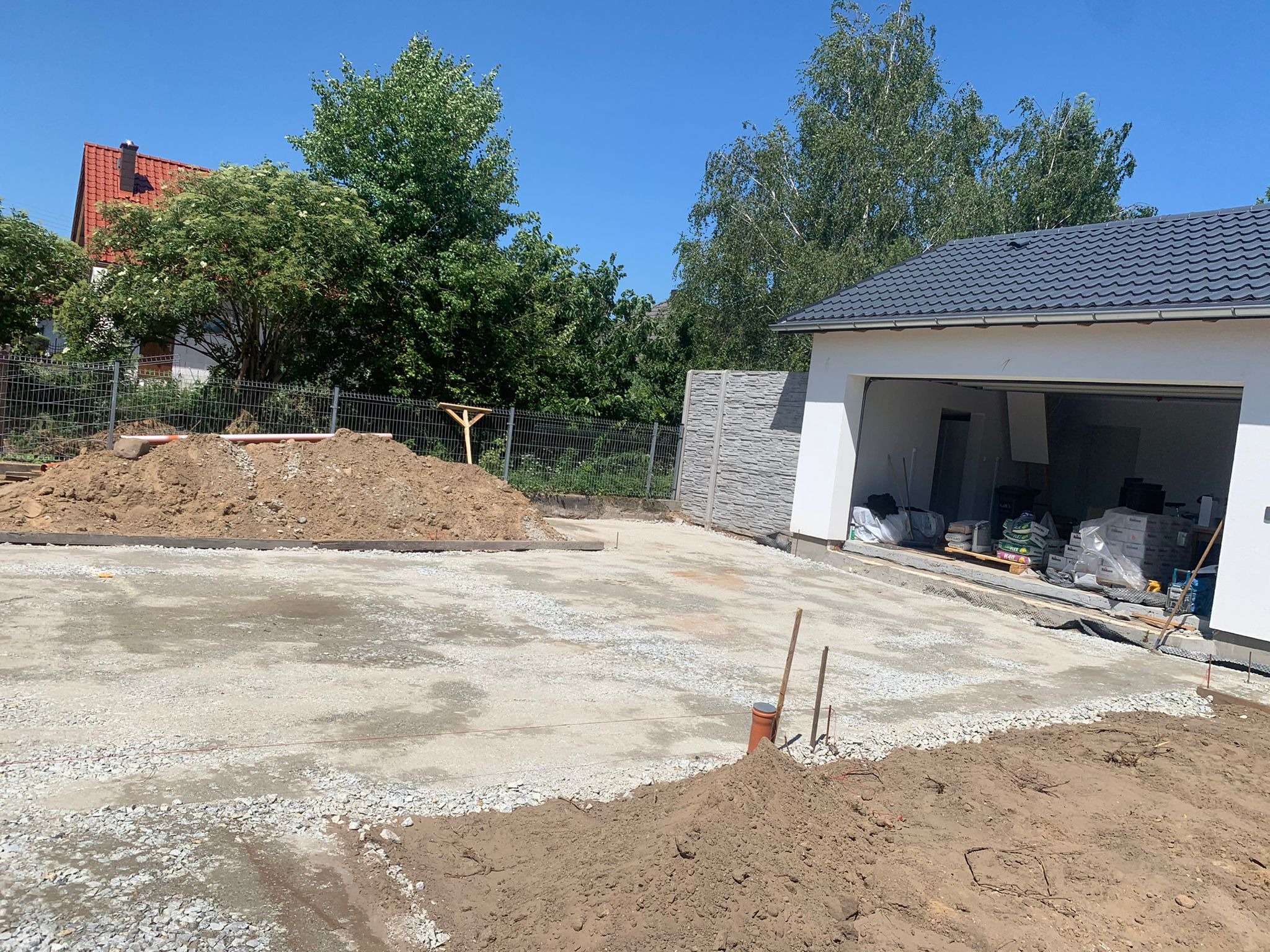 Przygotowany teren pod budowę podjazdu z widocznymi obrzeżami, nasypami ziemi oraz materiałami budowlanymi składowanymi w garażu.