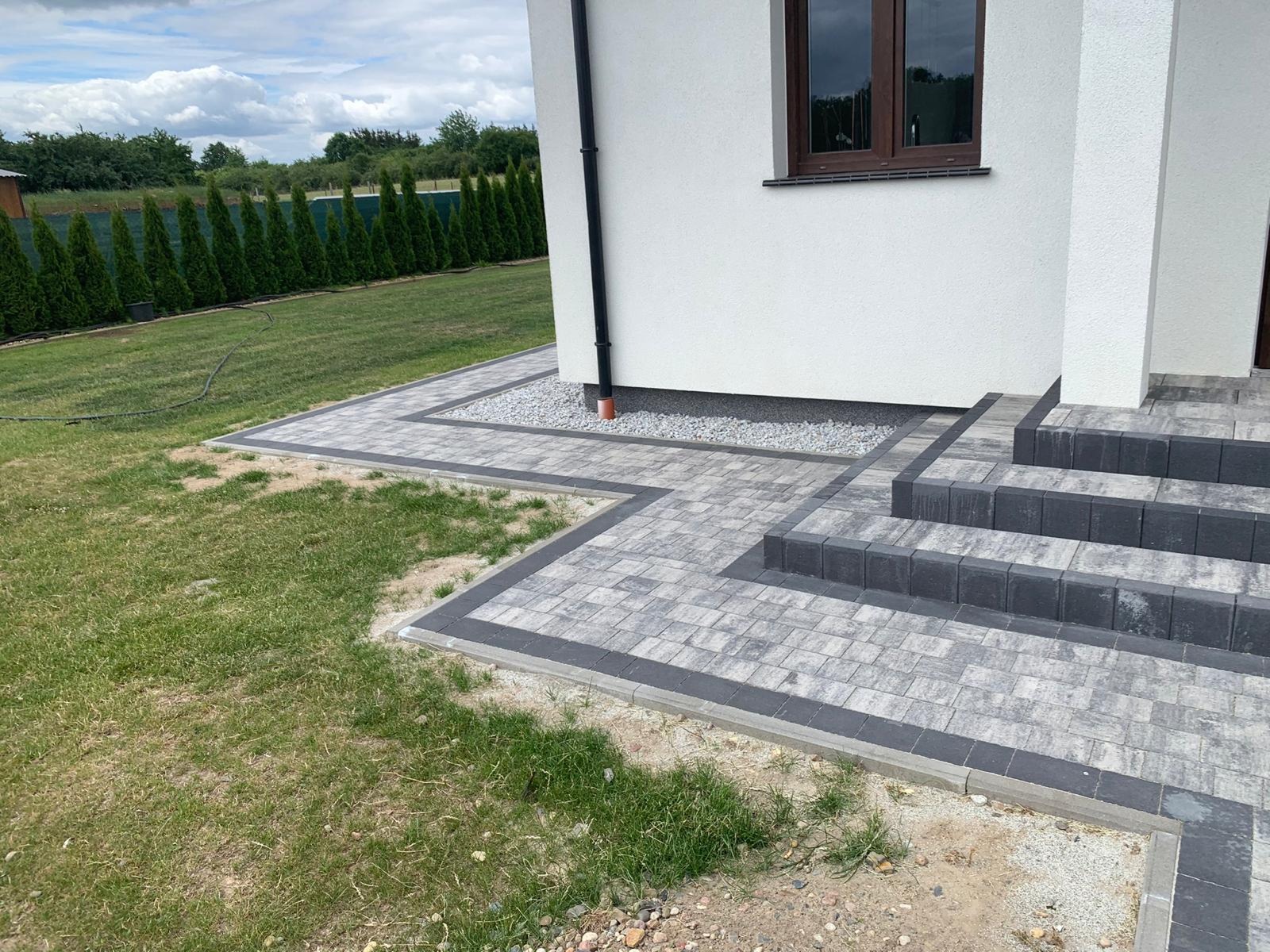 Szara kostka brukowa ułożona w geometryczny wzór wokół domu z białymi ścianami, widoczne schody wejściowe i pas żwiru przy ścianie, trawnik z tujami w tle.