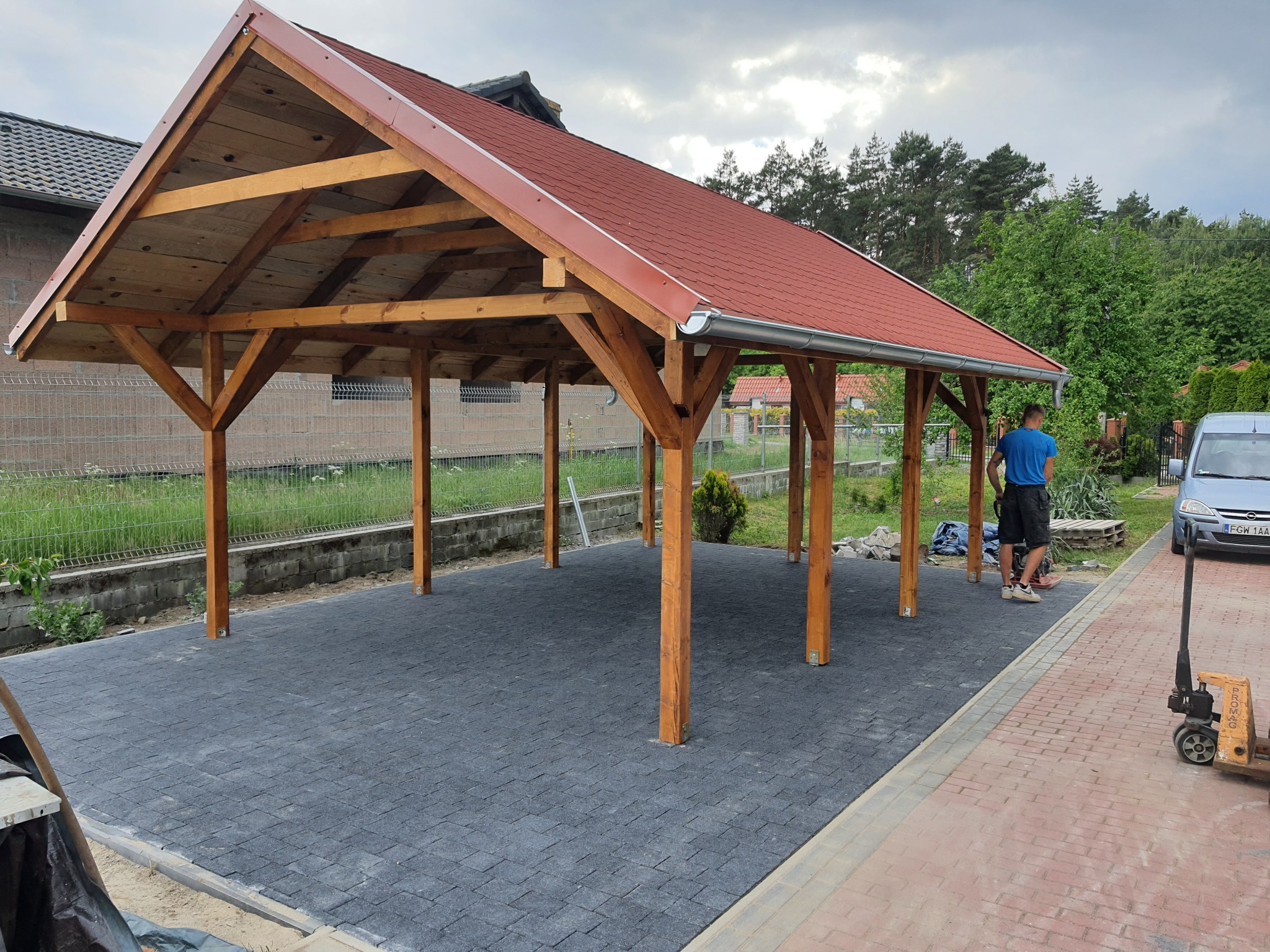 Zadaszona wiata garażowa z drewnianą konstrukcją i czerwoną dachówką, ułożona na szarej kostce brukowej, z widocznym fragmentem chodnika z czerwonej kostki, samochodem osobowym i pracownikiem w tle.