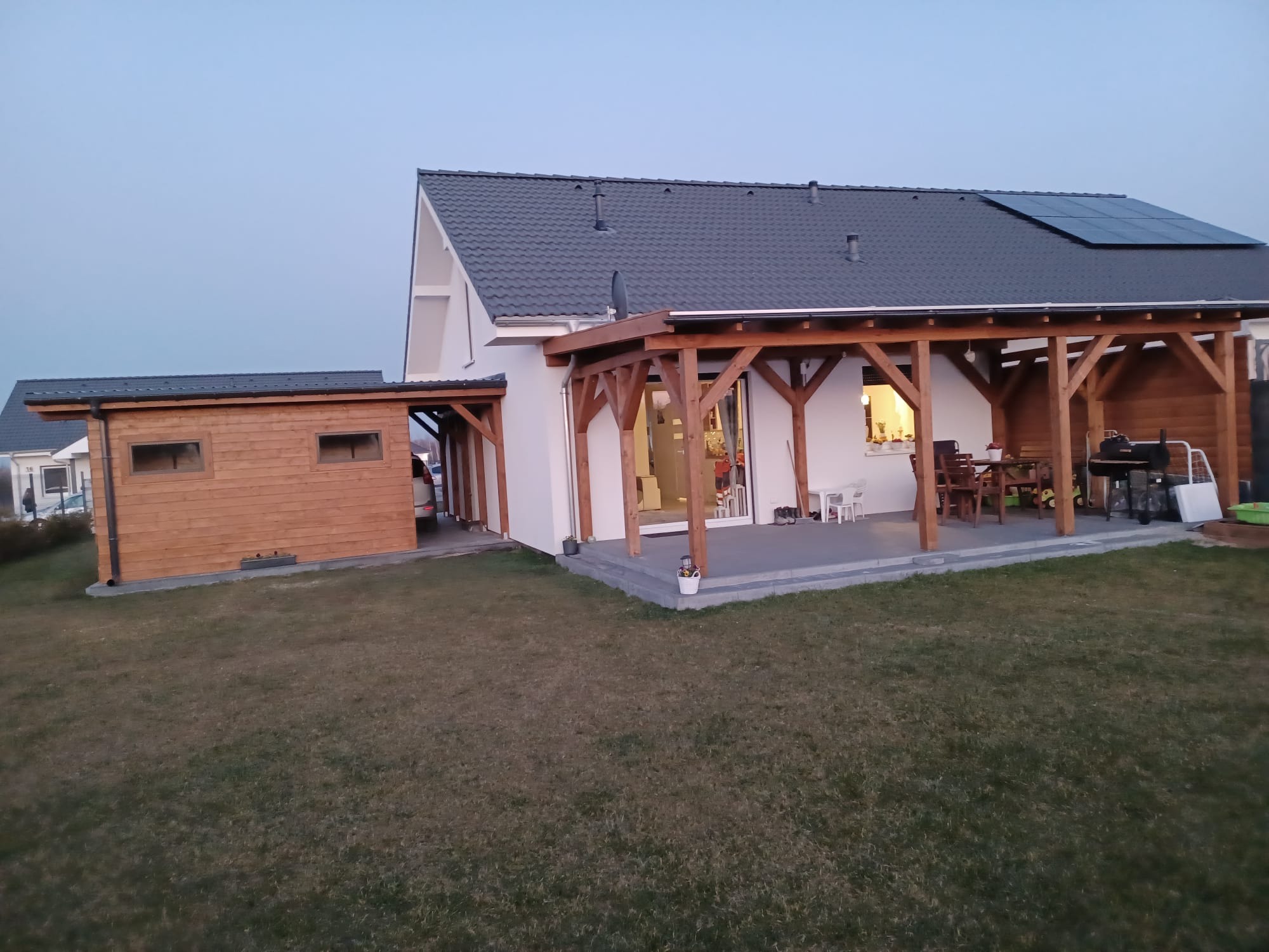 Nowoczesny dom jednorodzinny z drewnianą pergolą, tarasem wyłożonym szarą kostką i drewnianym budynkiem gospodarczym, widok z ogrodu o zmierzchu.