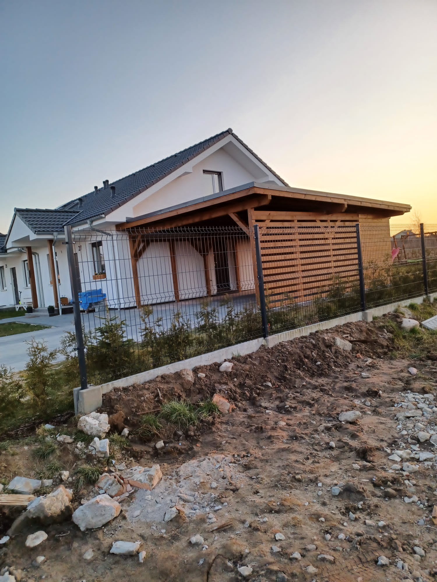 Nowoczesny dom jednorodzinny z drewnianą wiatą garażową i metalowym ogrodzeniem panelowym, widoczne ślady prac ziemnych w otoczeniu.