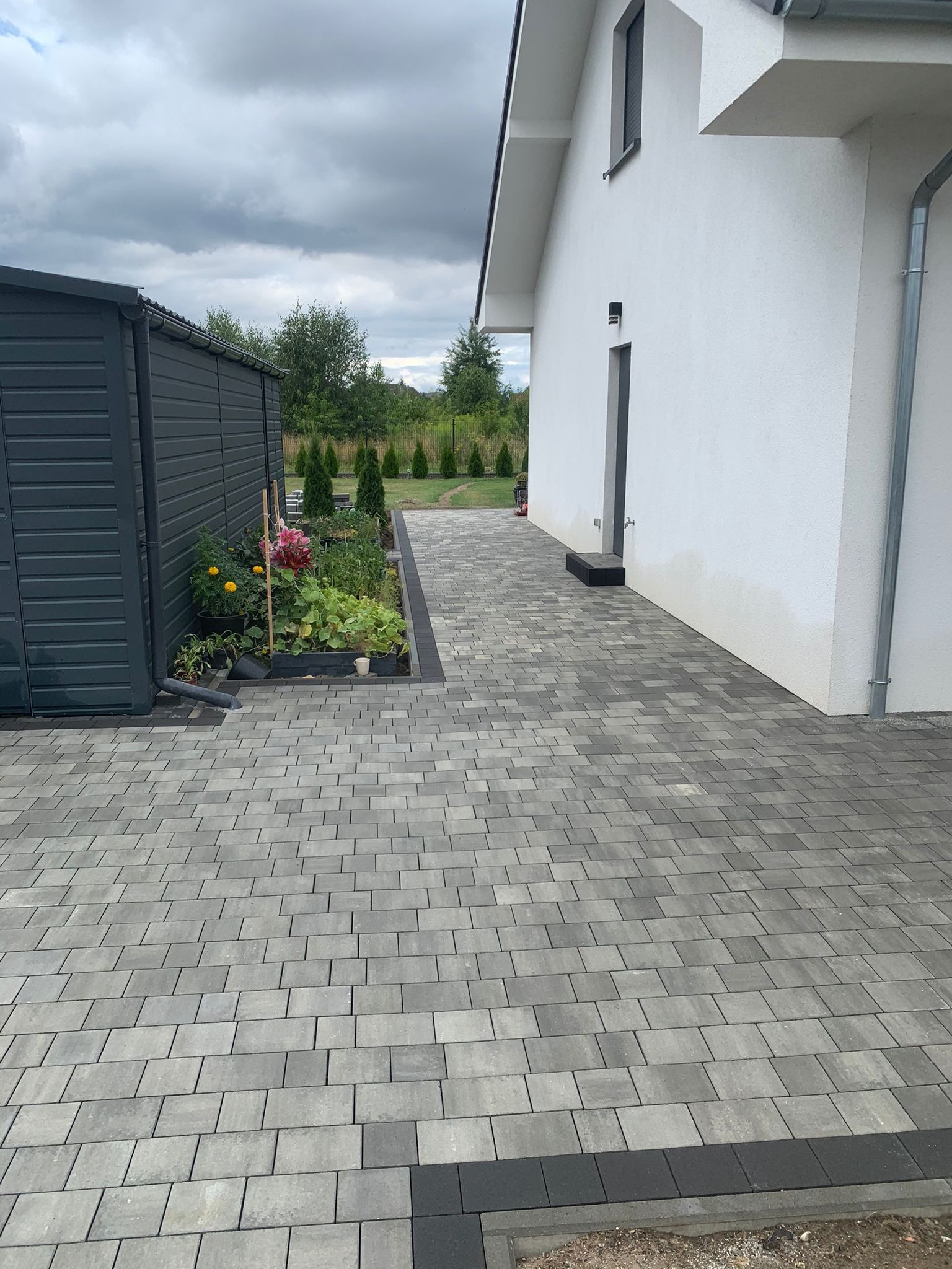 Szara kostka brukowa ułożona na podjeździe przed nowoczesnym, białym domem z szarą stolarką okienną i drzwiami, obok ciemnoszarej drewutni i rabaty z kwiatami.