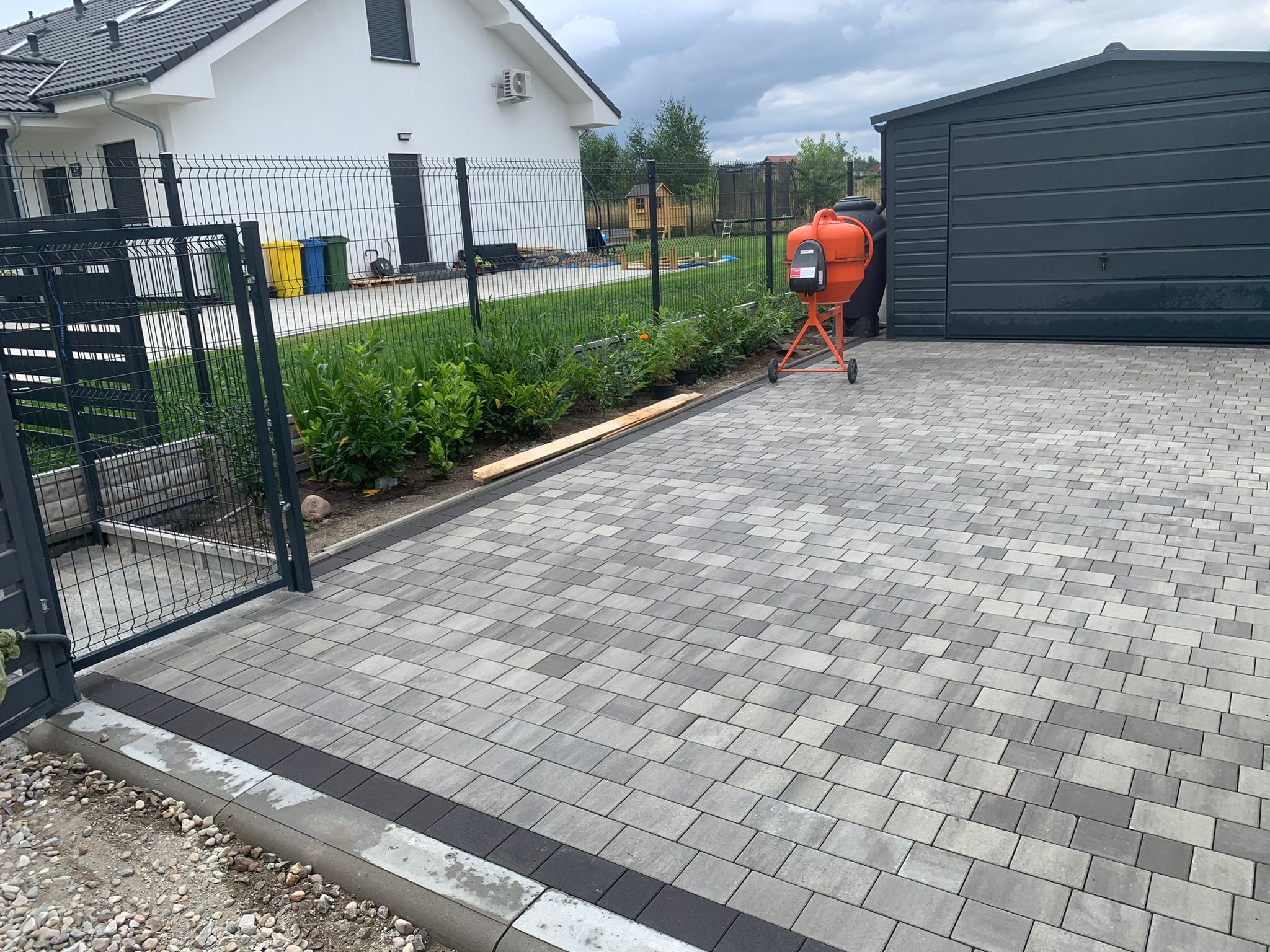 Ułożona szara kostka brukowa na podjeździe, z pomarańczową betoniarką w tle i elementami ogrodzenia posesji.