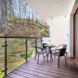 Zadaszony balkon z drewnianą podłogą, szklaną balustradą i widokiem na zalesione wzgórze. Cztery czarne krzesła z plecionki i okrągły stolik z dwiema filiżankami.