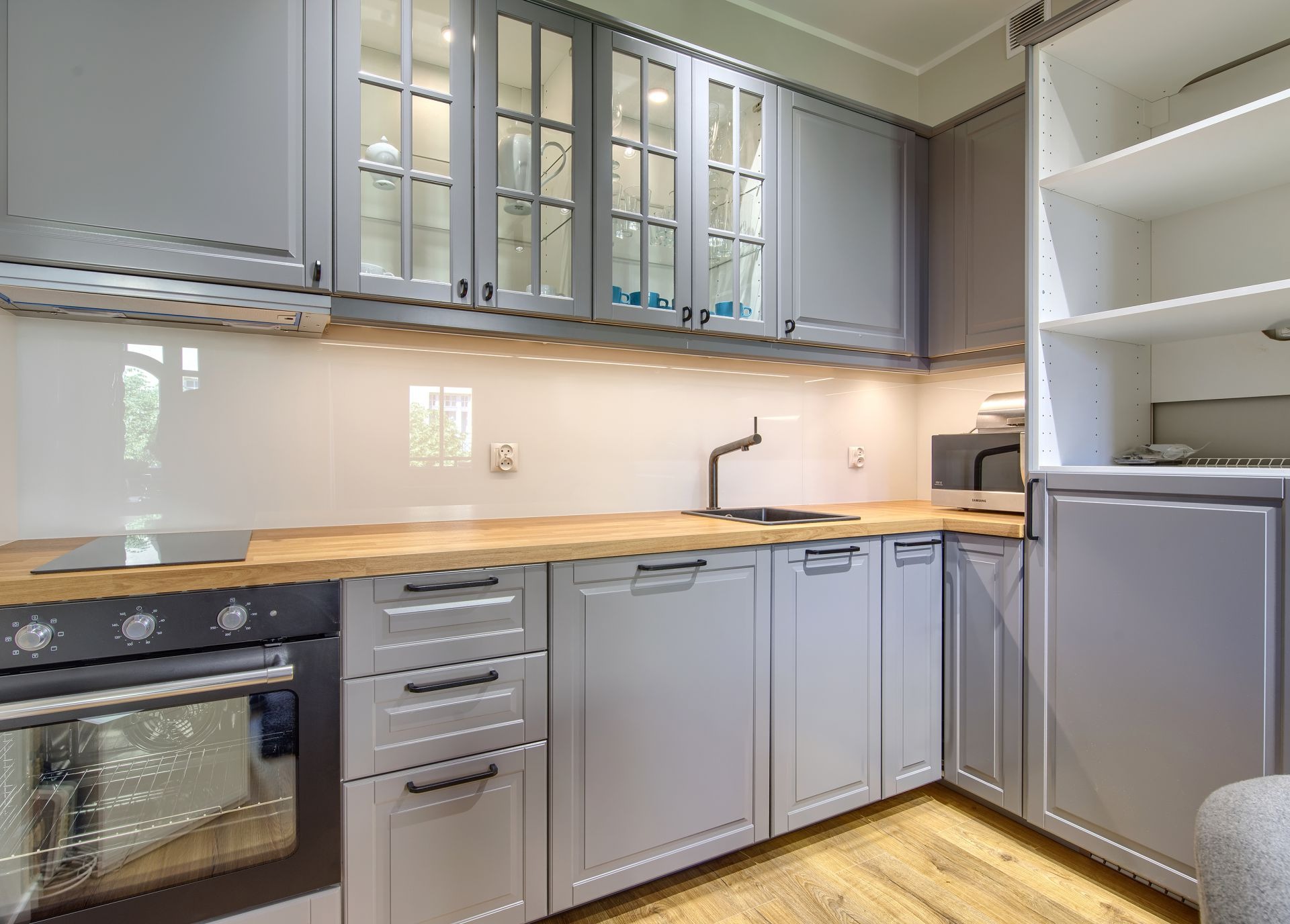 Szara kuchnia z drewnianym blatem, przeszklonymi szafkami górnymi z oświetleniem LED, czarną armaturą i parkietem na podłodze.