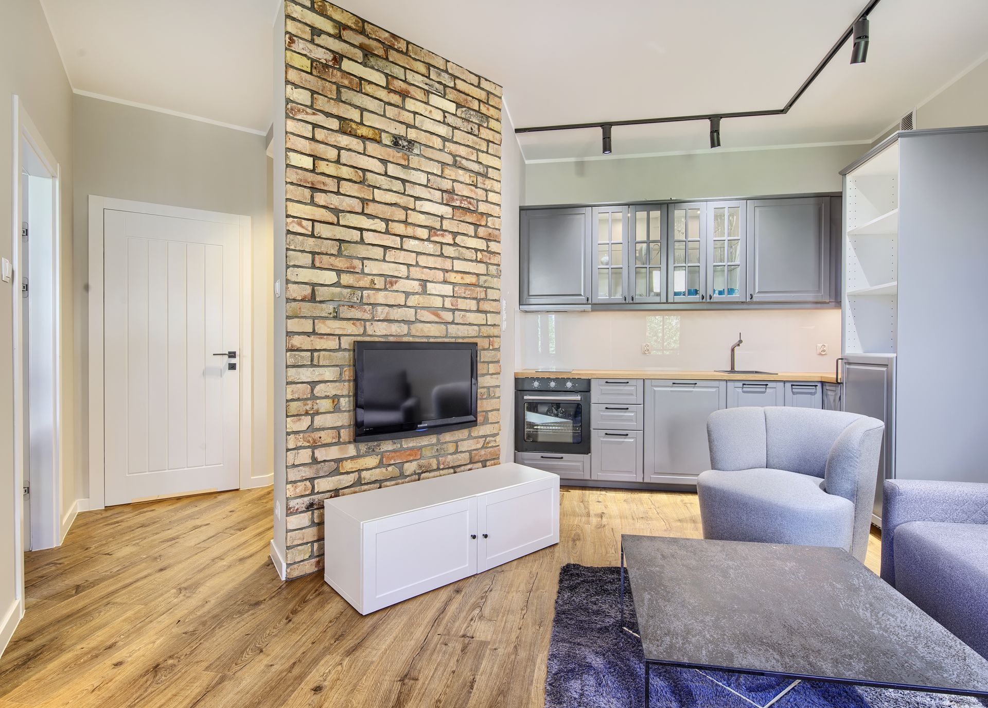 Salon z aneksem kuchennym, ściana z cegły dekoracyjnej, drewniana podłoga, szare meble kuchenne i wypoczynkowe, biała szafka RTV pod telewizorem.