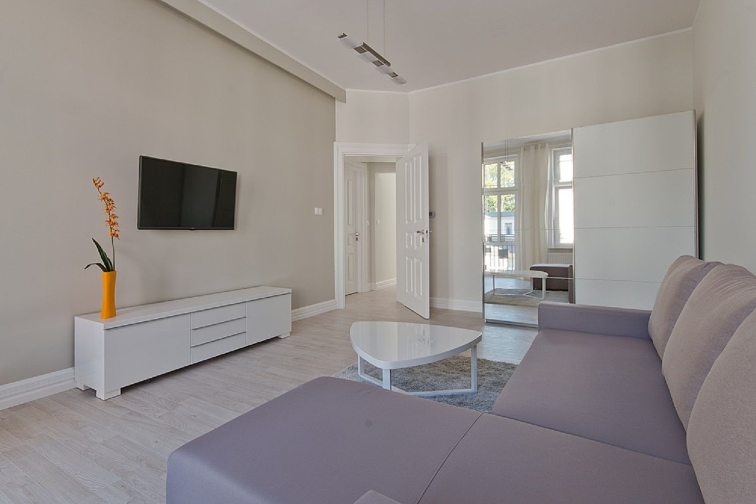 Jasne wnętrze salonu z szarą sofą narożną, białymi meblami, telewizorem na ścianie, lustrzaną szafą przesuwną i drewnianą podłogą w odcieniach szarości.