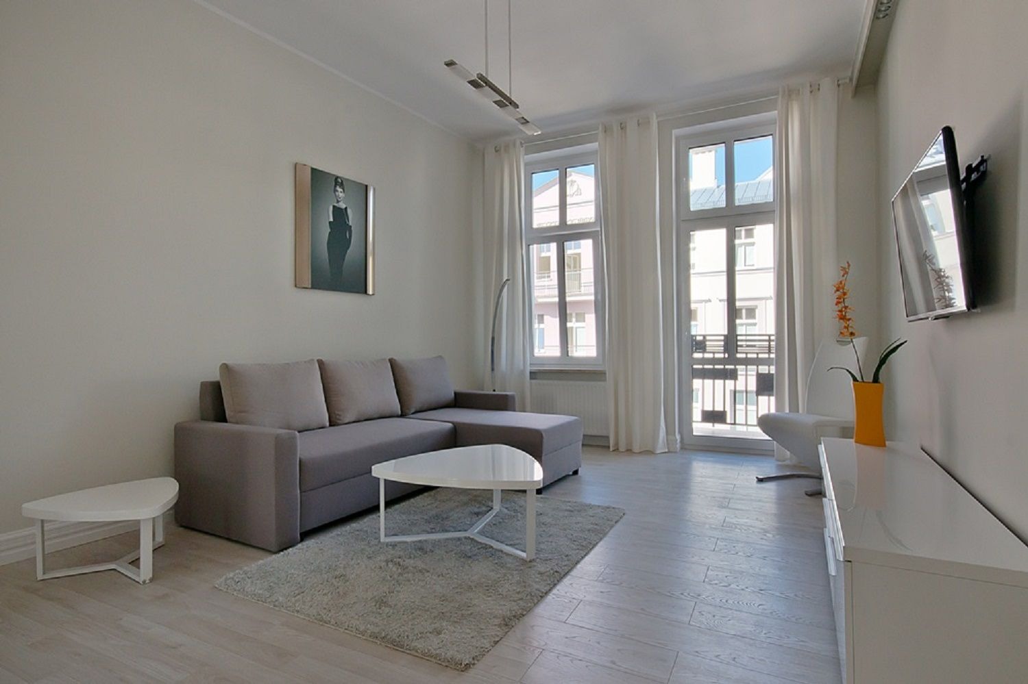 Jasne wnętrze salonu z szarą sofą narożną, białym stolikiem kawowym o trójkątnym blacie, telewizorem na ścianie i parkietem w odcieniach szarości, uzupełnione o białe zasłony i obraz w ramie.