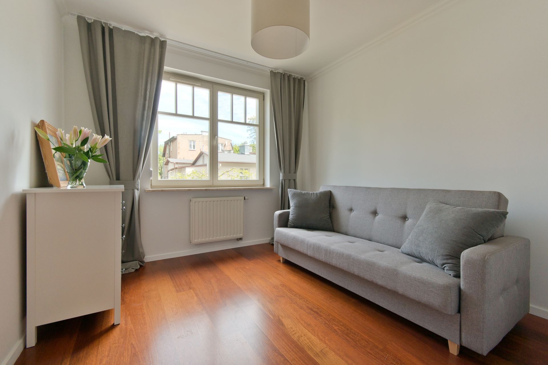 Jasne wnętrze pokoju z drewnianą podłogą, szarą sofą z poduszkami, komodą z kwiatami w wazonie i oknem z szarymi zasłonami.