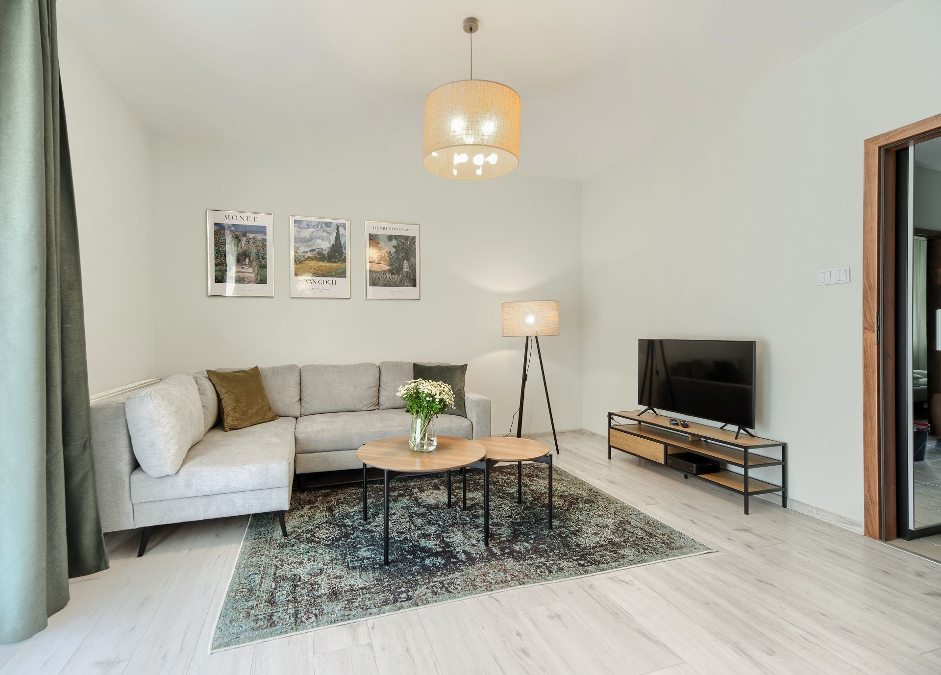 Jasne wnętrze salonu z narożną sofą, okrągłymi stolikami kawowymi, dywanem w stylu vintage i drewnianą podłogą. Na ścianie wiszą trzy plakaty z motywami pejzaży. W tle widoczny telewizor na niskiej...