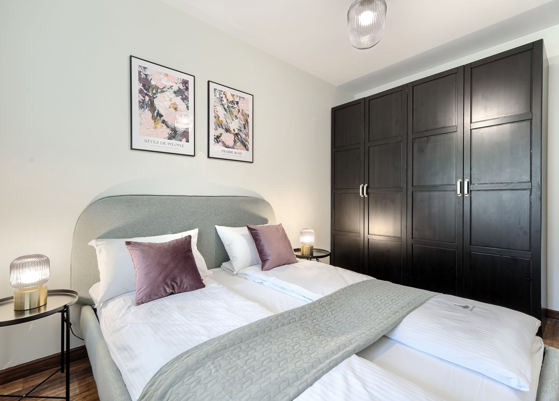Nowoczesna sypialnia z łóżkiem małżeńskim, szarą tapicerowaną ramą, ciemną szafą z czterema drzwiami, dwiema lampkami nocnymi i dekoracyjnymi obrazami na ścianie.