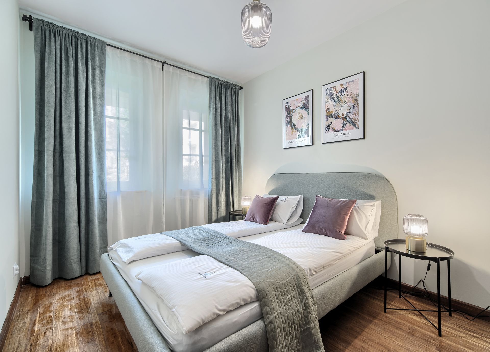 Nowoczesna sypialnia z łóżkiem kontynentalnym, drewnianą podłogą, szarymi zasłonami i lampkami nocnymi na metalowych stolikach.
