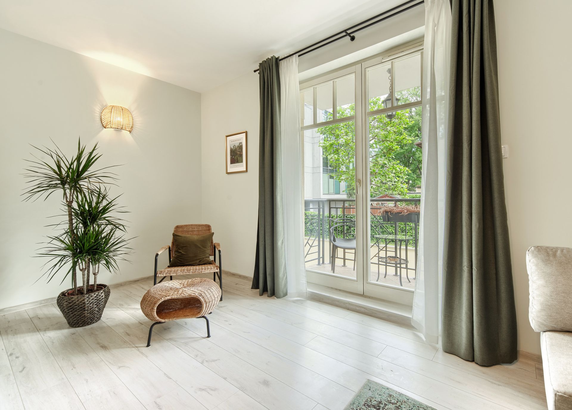 Jasne wnętrze pokoju z białymi panelami podłogowymi, wiklinowym fotelem z poduszką, rośliną doniczkową, wiklinowym stolikiem i dużym oknem balkonowym z szarymi i białymi zasłonami.