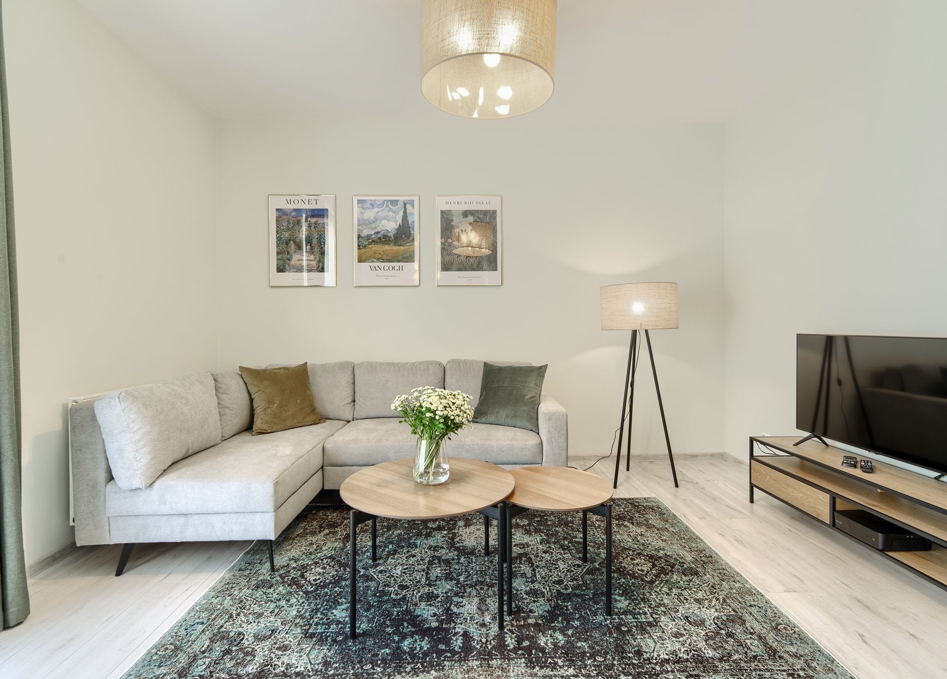 Jasny salon z narożną kanapą, dywanem w stylu vintage, okrągłymi stolikami kawowymi, lampą podłogową na trójnogu i obrazami w ramach na ścianie.