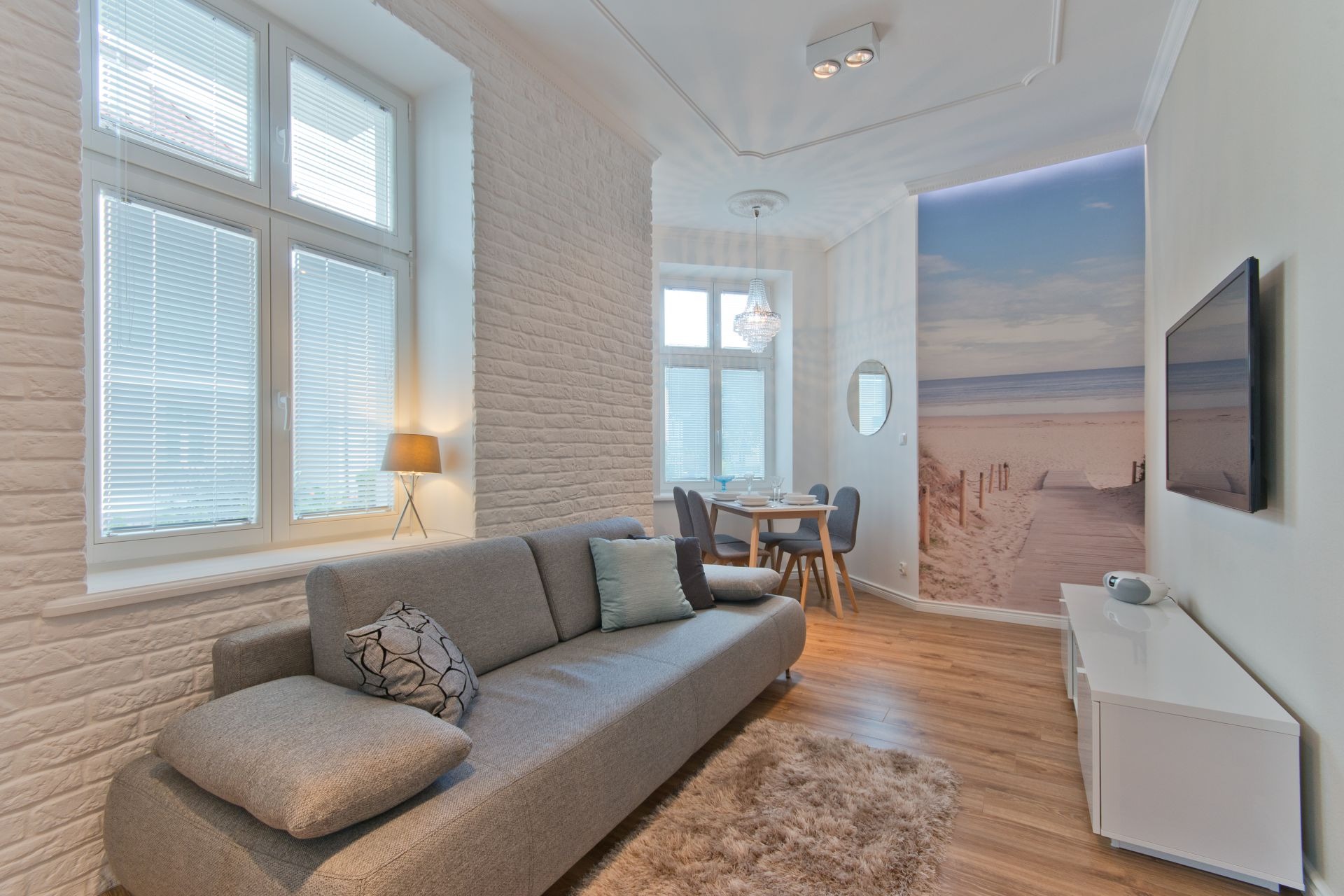 Jasne wnętrze pokoju dziennego z szarą sofą, ceglaną ścianą pomalowaną na biało, oknami z żaluzjami, fototapetą z motywem plaży oraz drewnianą podłogą.
