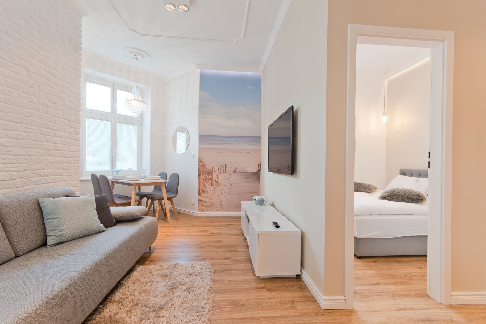 Jasne, nowocześnie urządzone wnętrze mieszkania z drewnianą podłogą, białymi ścianami z cegły, szarą sofą, stołem z krzesłami, telewizorem, szafką RTV i fototapetą z motywem plaży.