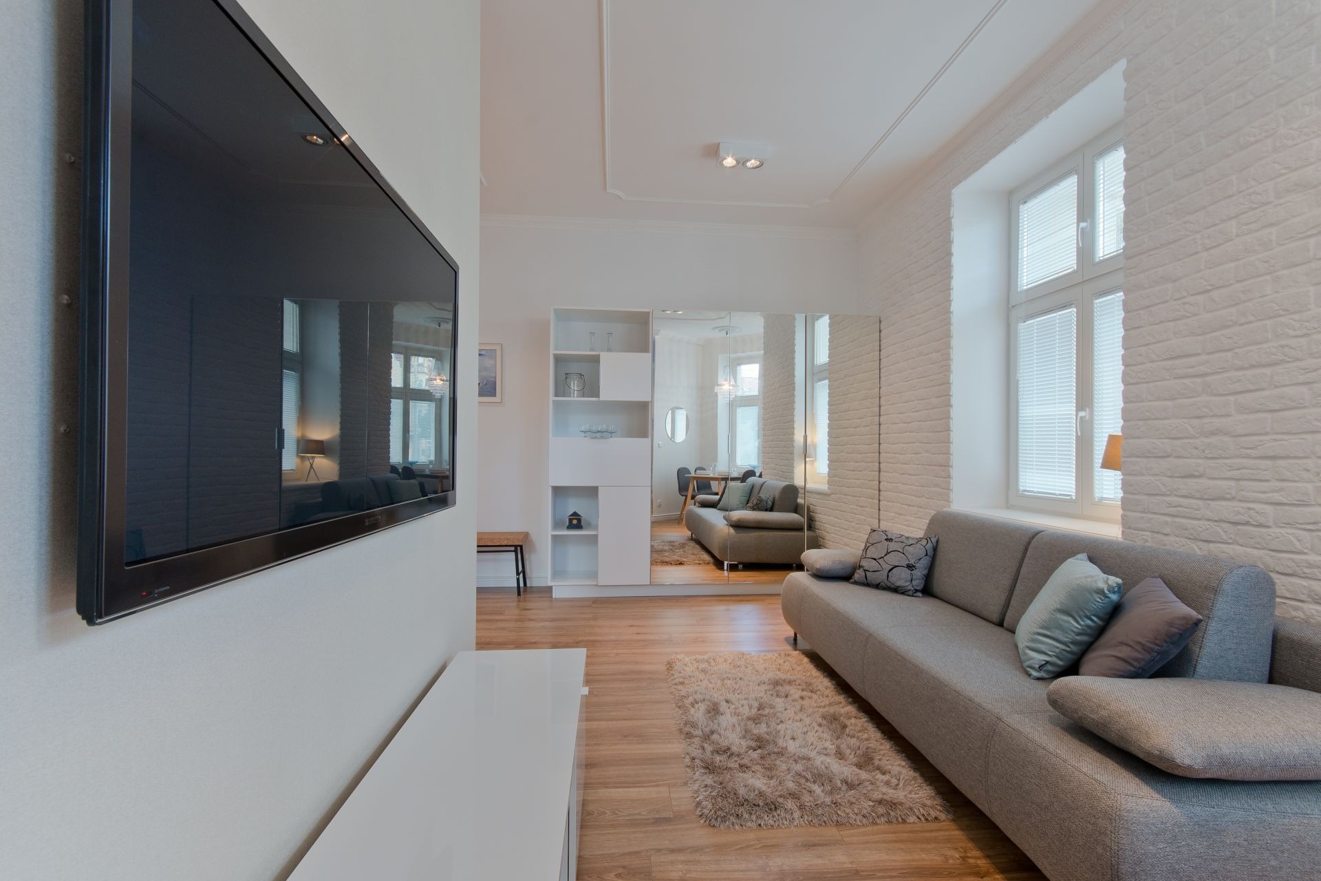 Jasne wnętrze salonu z telewizorem na ścianie, szarą sofą z poduszkami, białymi cegłami na ścianie i dużym lustrem w tle.