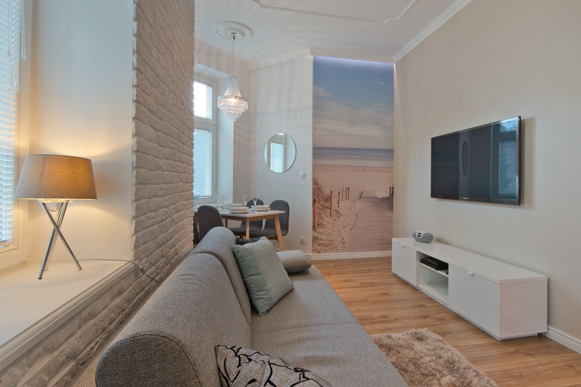 Jasne wnętrze salonu z ceglaną kolumną, lampą na parapecie, szarą sofą, stołem jadalnym z krzesłami, telewizorem na ścianie i fototapetą z motywem plaży.