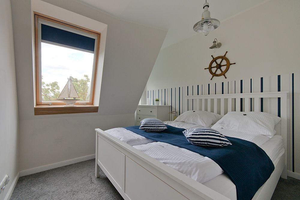 Pokój w stylu marynistycznym z białym łóżkiem, drewnianym oknem dachowym i dekoracjami: koło sterowe na ścianie oraz model żaglówki na parapecie.