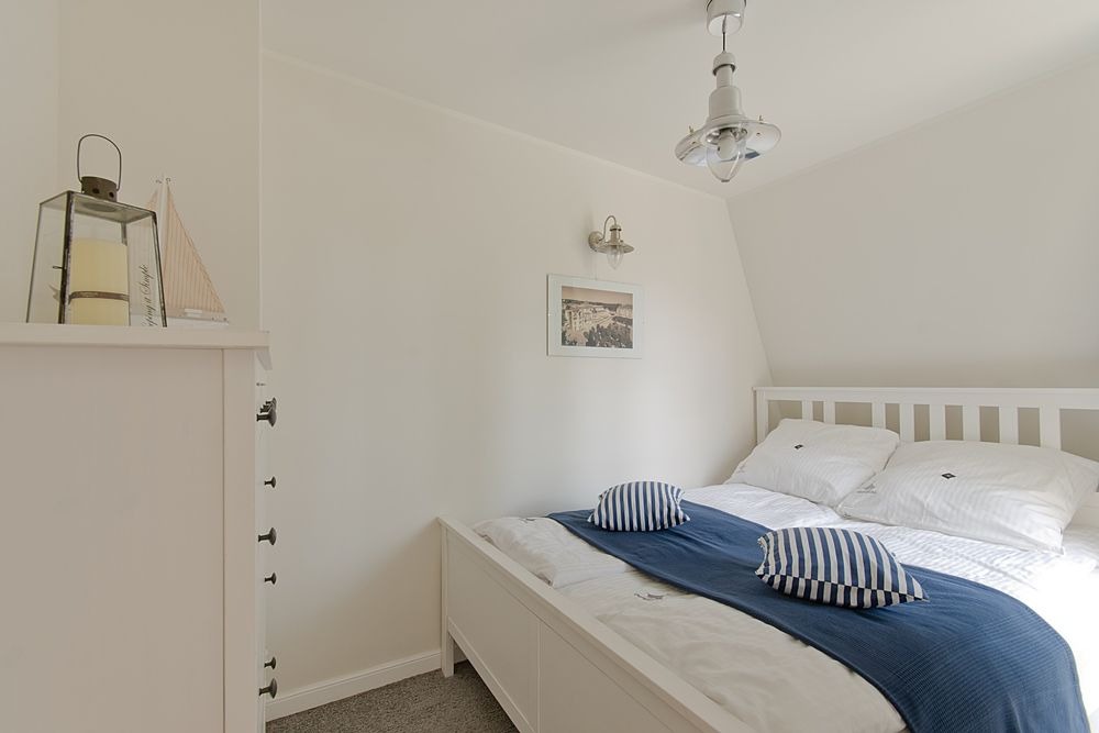 Jasna sypialnia w stylu marynistycznym z białym łóżkiem, niebieską narzutą i poduszkami w paski, widoczna lampa sufitowa w stylu retro i dekoracje: lampion oraz model żaglówki na komodzie.