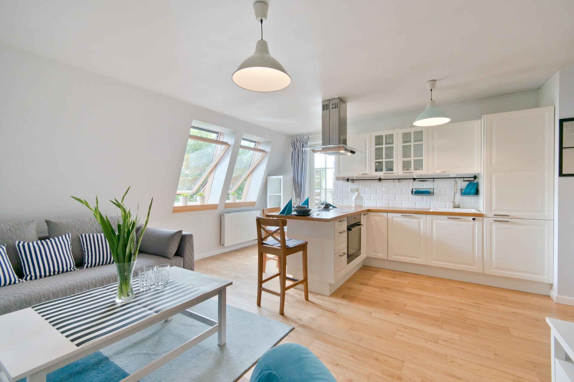 Jasne wnętrze z aneksem kuchennym: białe szafki, drewniany blat, wyspa z krzesłami barowymi w paski, widok na okna dachowe i salon z szarą sofą w paski.