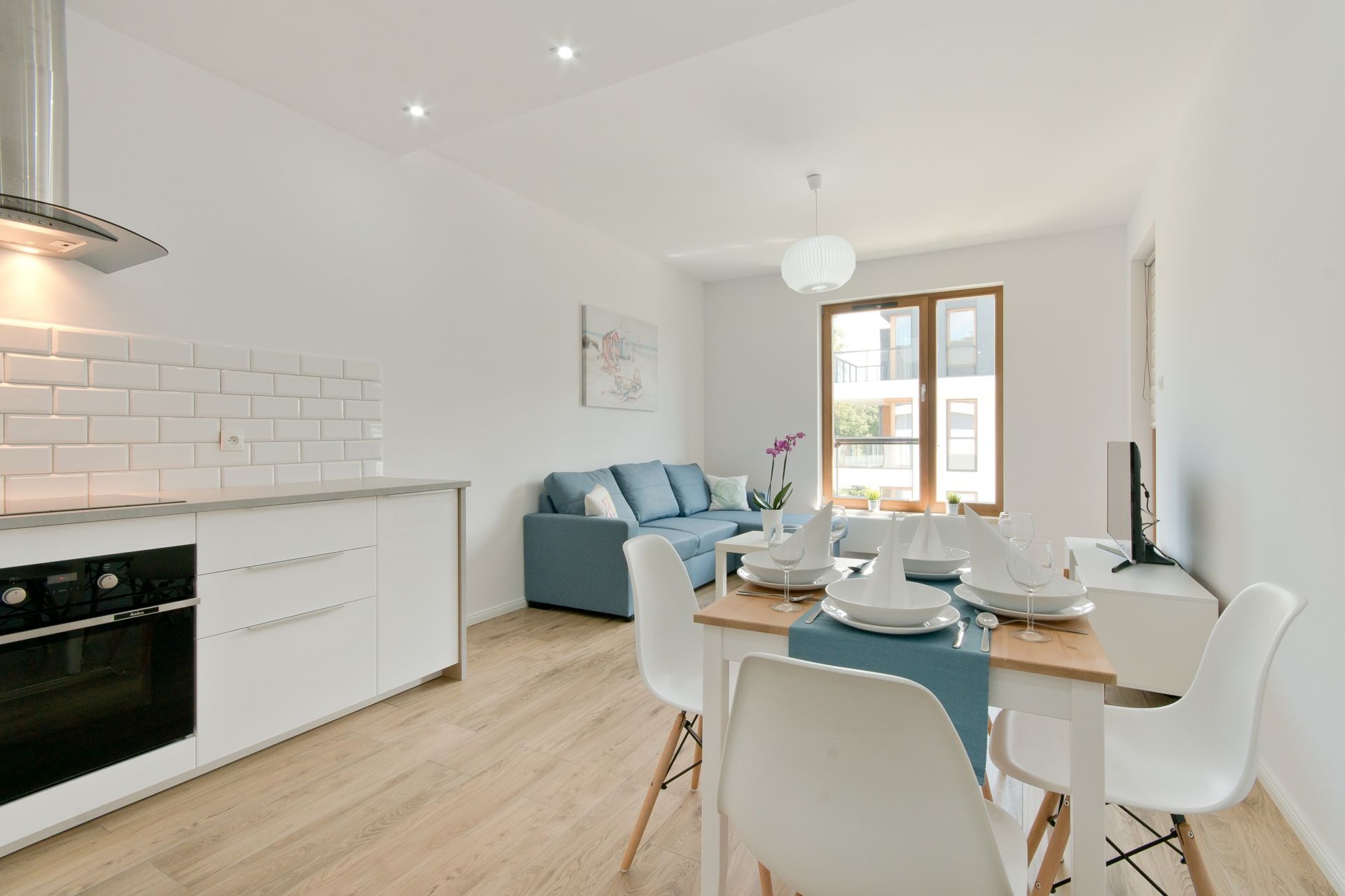 Jasne, przestronne wnętrze mieszkania z aneksem kuchennym, stołem jadalnym i sofą w odcieniach błękitu. Minimalistyczny wystrój z białymi meblami i drewnianą podłogą.
