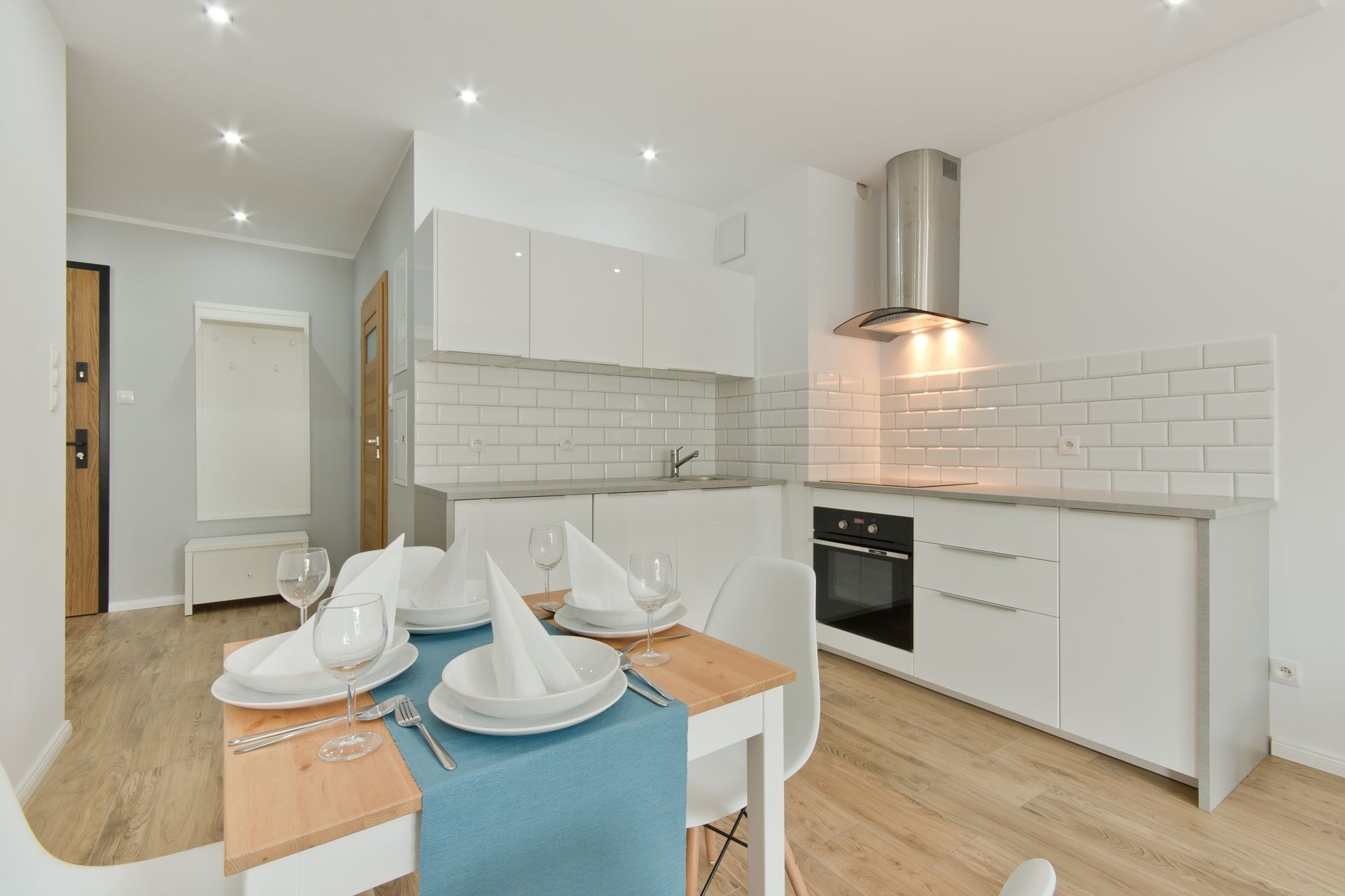 Nowoczesna, jasna kuchnia z białymi meblami, białymi płytkami na ścianie i drewnianą podłogą, nakryty stół z niebieskim bieżnikiem i białymi naczyniami, widok z perspektywy stołu.